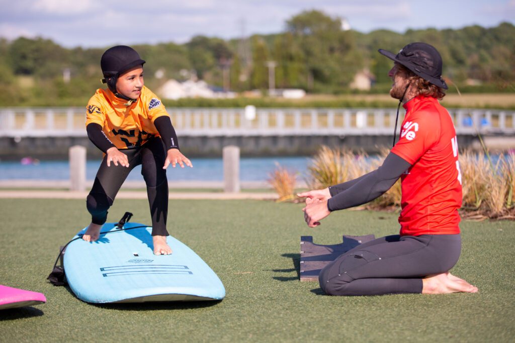 A child stands on a surfboard on land, in a lesson with a coach at The Wave Bristol.